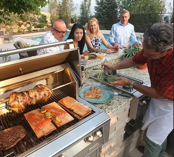 A built-in outdoor kitchen by a pool, with salmon and chicken on the grill and guests standing at the counter.